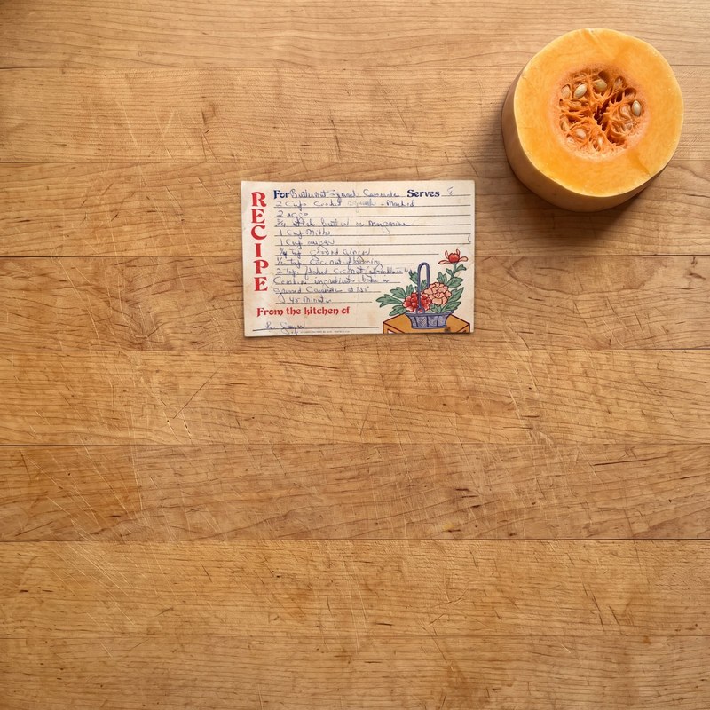 Before: the original handwritten Butternut Squash Casserole card lying on a maple cutting board beside a slice of butternut squash.